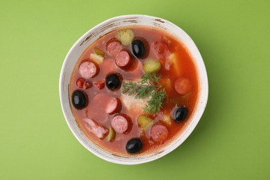 Photo of Meat solyanka soup with thin dry smoked sausages in bowl on light green background, top view