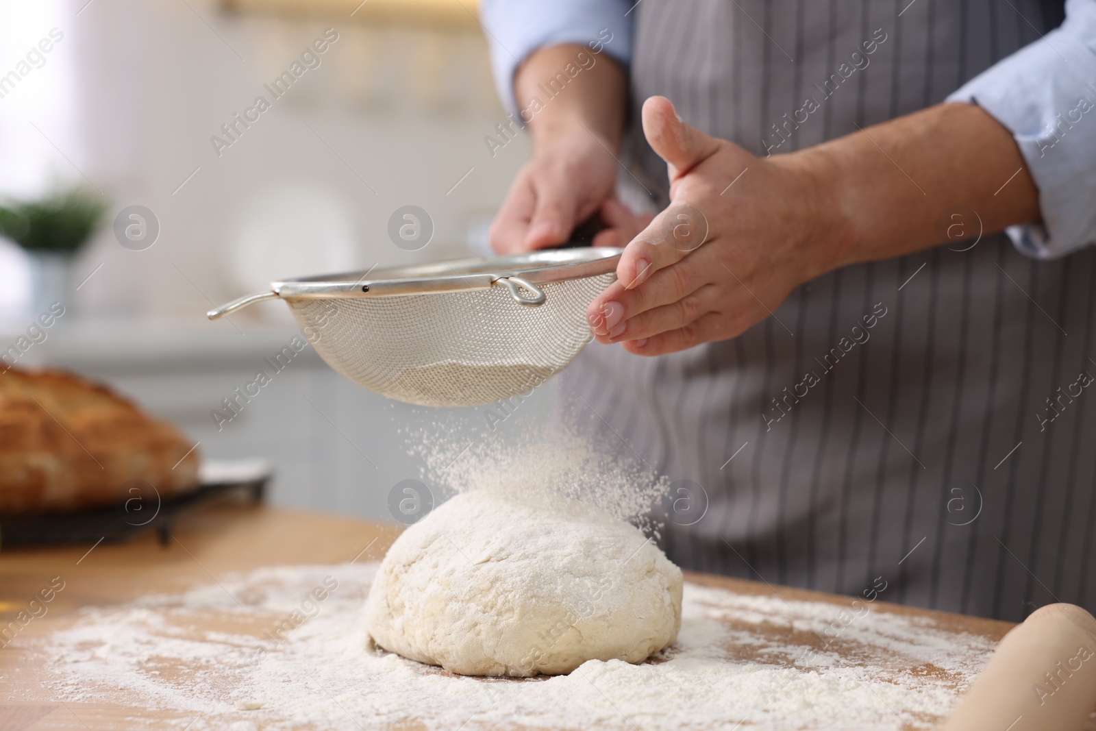 Photo of Making bread. Man sprinkling flour onto dough at wooden table in kitchen, closeup