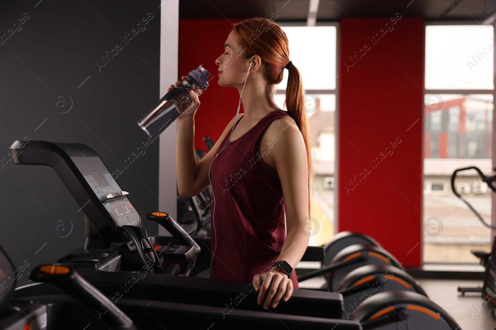 Photo of Athletic young woman with earphones and bottle of water training on treadmill in gym