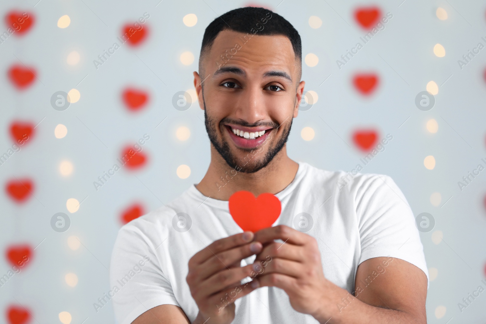 Photo of Handsome young man with red paper heart indoors, view from camera. Valentine's day celebration in long distance relationship