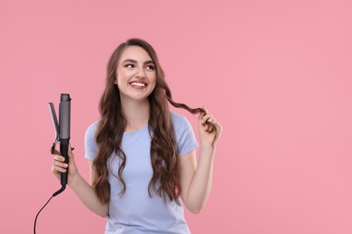 Photo of Happy young woman with beautiful hair holding curling iron on pink background, space for text