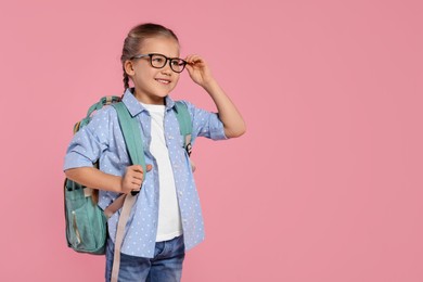 Happy schoolgirl in glasses with backpack on pink background, space for text