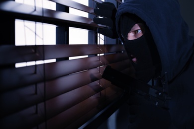 Masked man with gun spying through window blinds indoors. Criminal offence