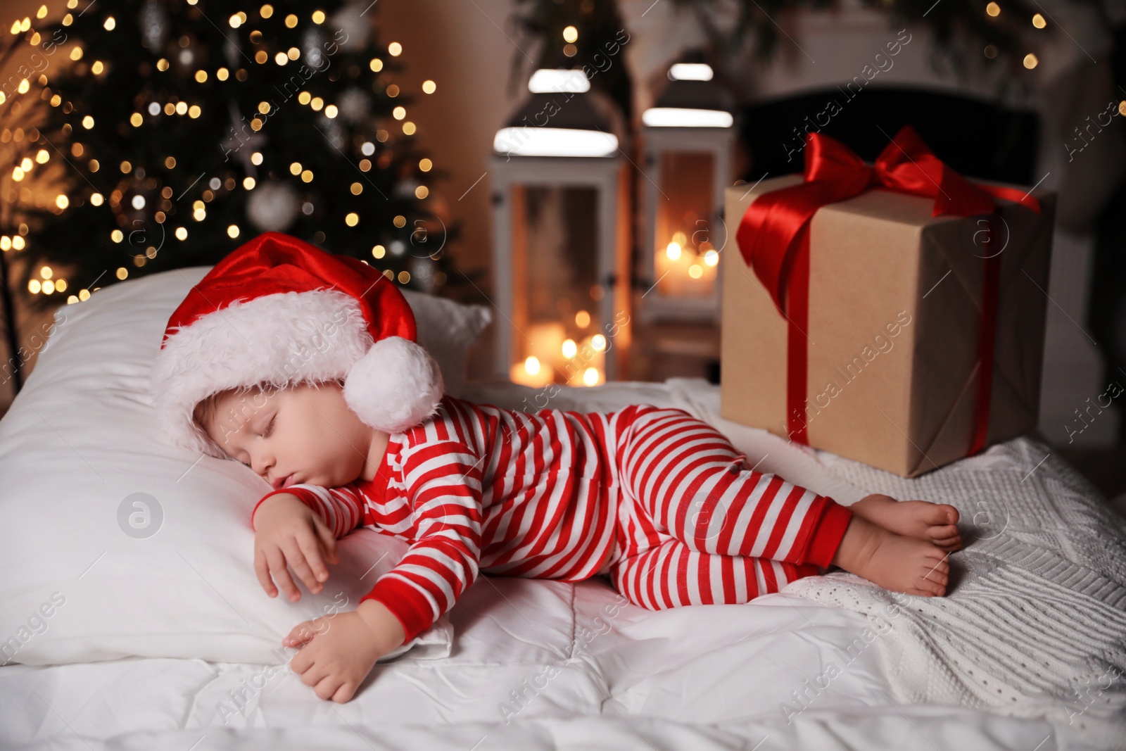 Photo of Baby in Christmas pajamas and Santa hat sleeping near gift box on bed indoors