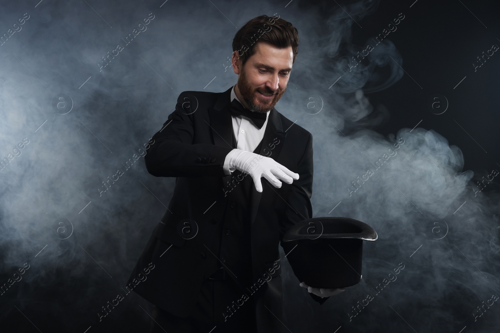 Photo of Happy magician showing magic trick with top hat in smoke on dark background