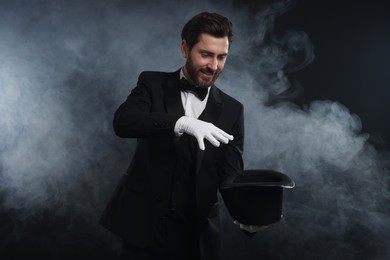 Happy magician showing magic trick with top hat in smoke on dark background