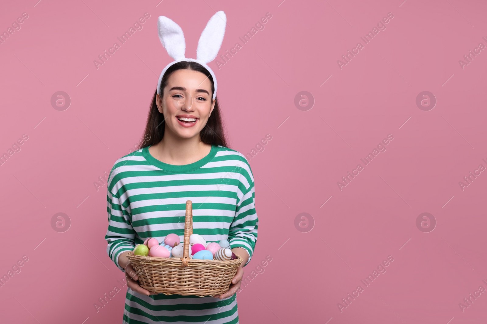 Photo of Happy woman in bunny ears headband holding wicker basket of painted Easter eggs on pink background. Space for text