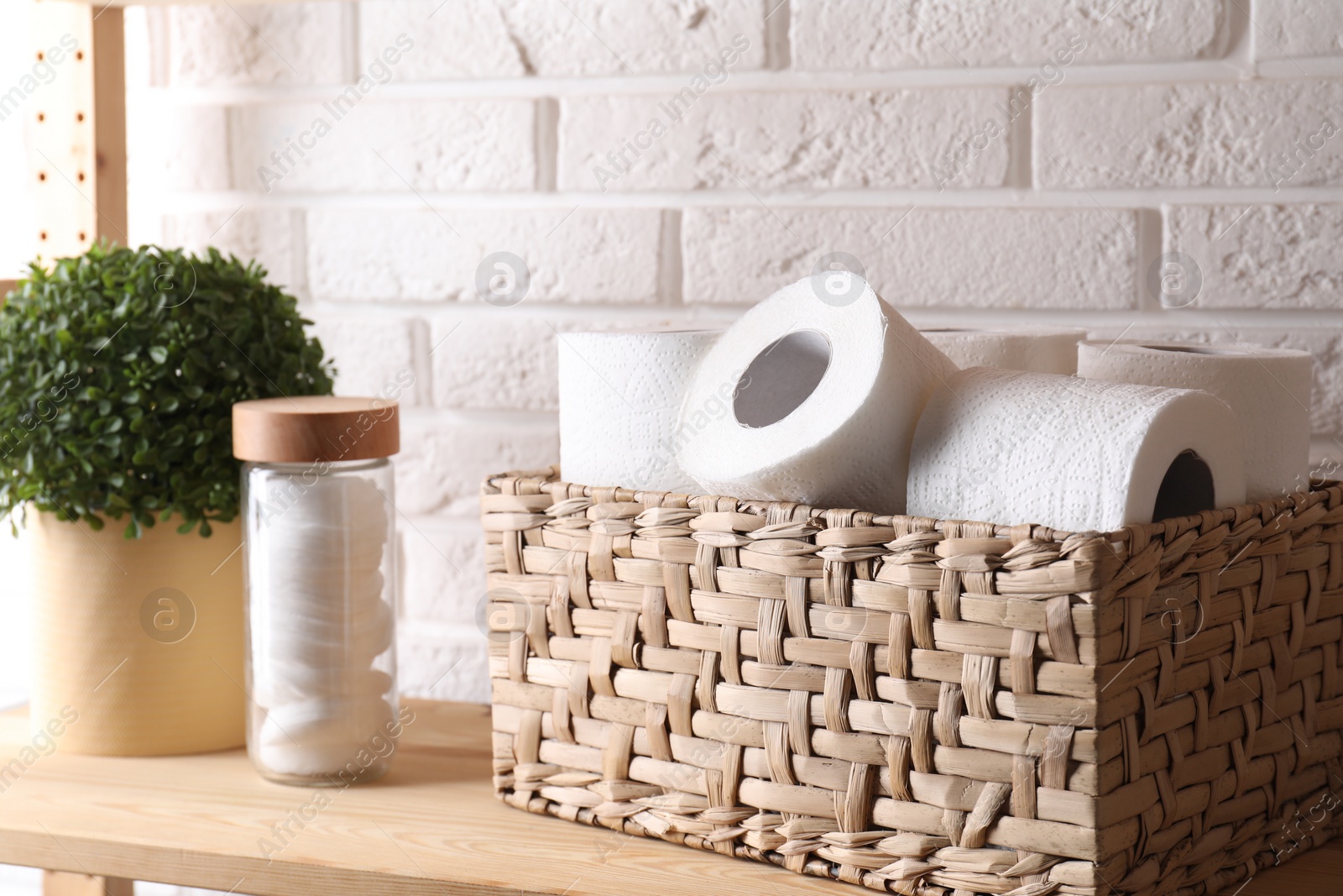 Photo of Toilet paper rolls in wicker basket, floral decor and cotton pads on wooden shelf near white brick wall