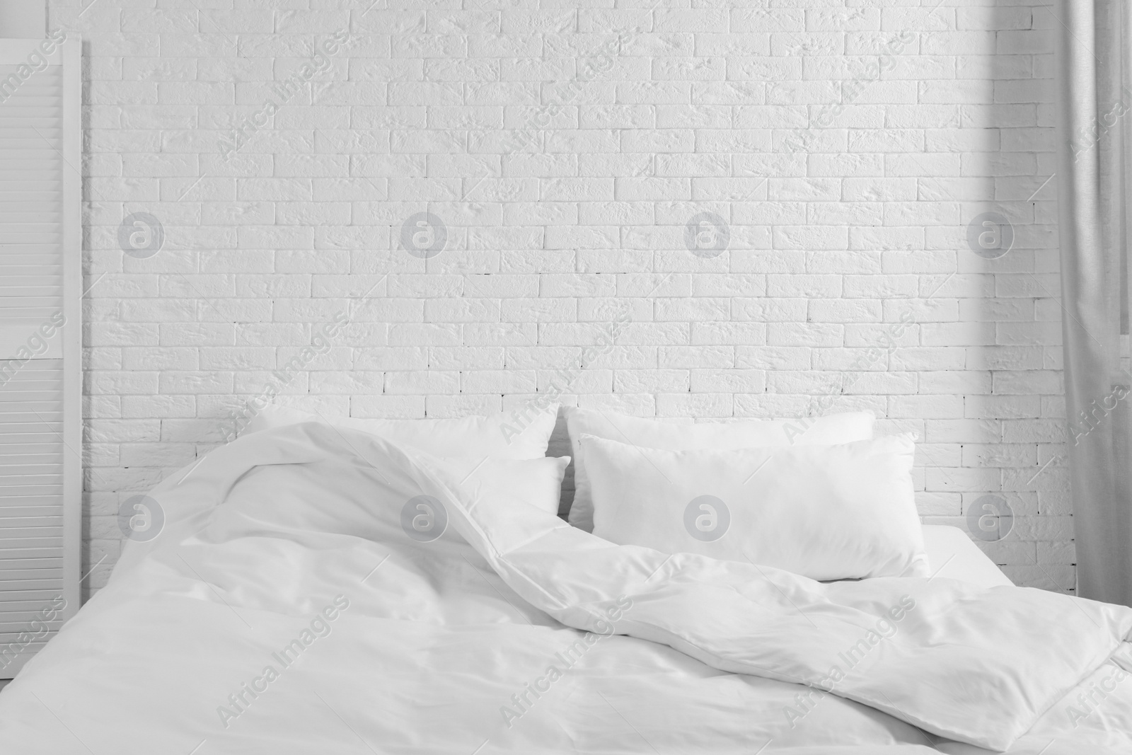 Photo of Large comfortable bed with pillows and blanket near white brick wall indoors. Stylish interior