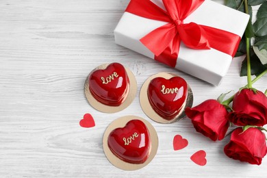 St. Valentine's Day. Delicious heart shaped cakes, roses and gift on white wooden table, flat lay. Space for text