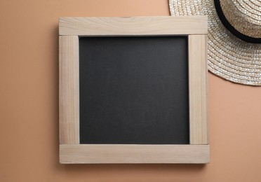 Photo of Blank chalkboard with straw hat on light brown background, flat lay. Space for text