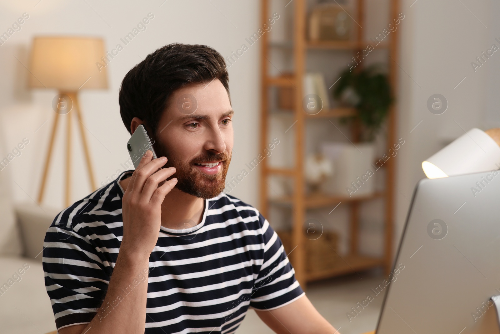 Photo of Home workplace. Happy man talking on smartphone while working with computer in room