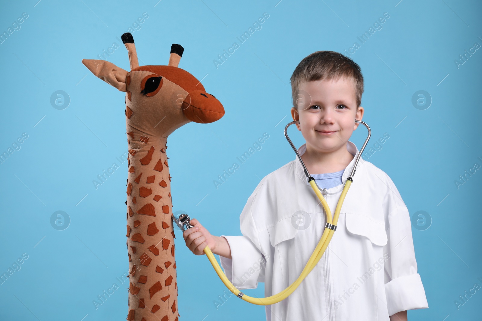 Photo of Cute little boy in pediatrician's uniform playing with stethoscope and toy giraffe on light blue background