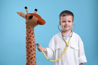 Photo of Cute little boy in pediatrician's uniform playing with stethoscope and toy giraffe on light blue background