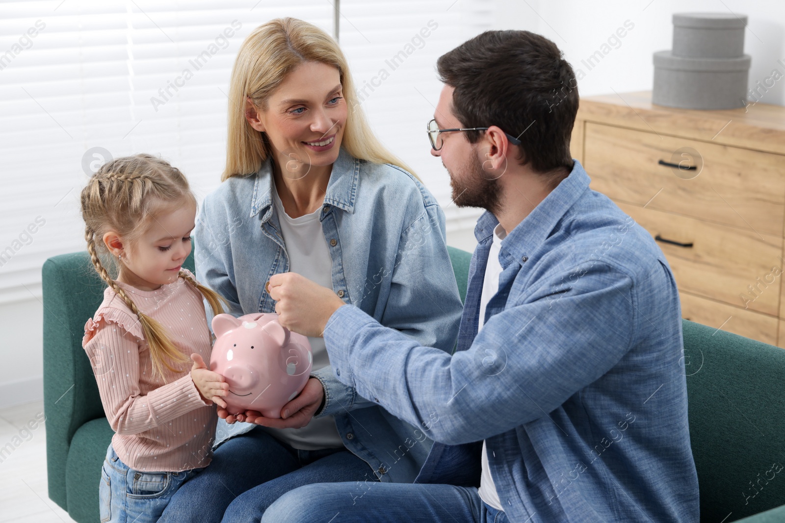 Photo of Planning budget together. Little girl with her parents putting coin into piggybank at home