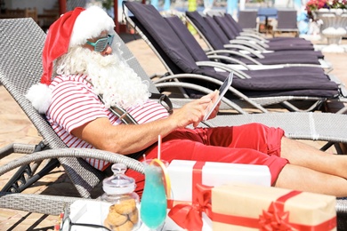 Photo of Authentic Santa Claus using tablet on lounge chair at resort