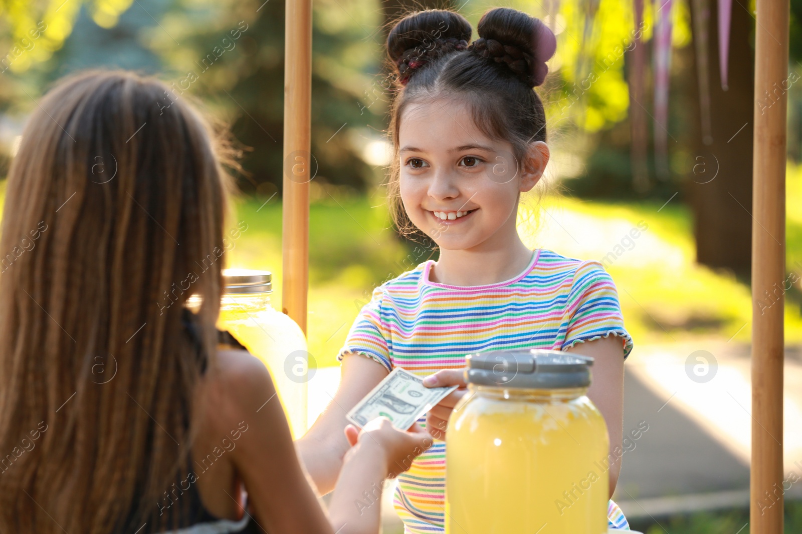 Photo of Little girl selling natural lemonade to kid in park. Summer refreshing drink