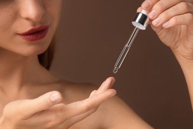 Beautiful young woman applying cosmetic serum onto her finger on brown background, closeup