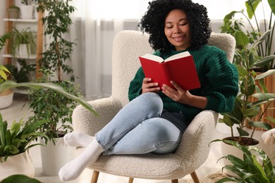 Relaxing atmosphere. Happy woman reading book in armchair surrounded by beautiful houseplants in room