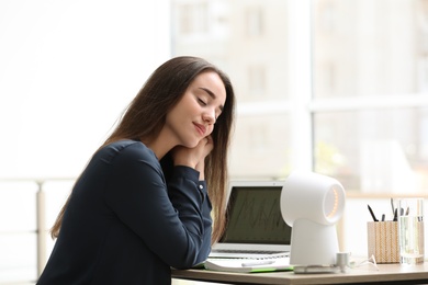 Young woman enjoying air flow from portable fan at workplace. Summer heat