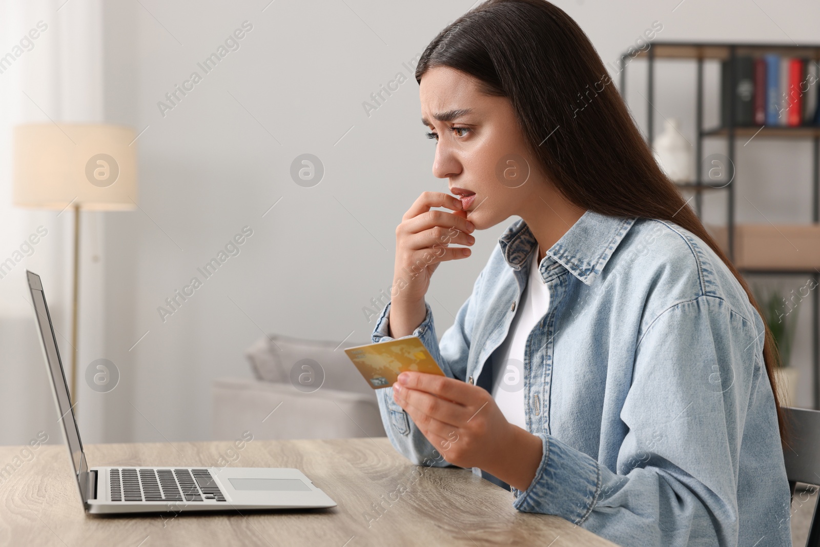 Photo of Stressed woman with credit card using laptop at table indoors. Be careful - fraud