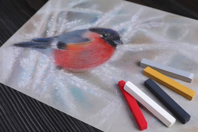 Photo of Colorful chalk pastels and beautiful painting of bullfinch on black wooden table