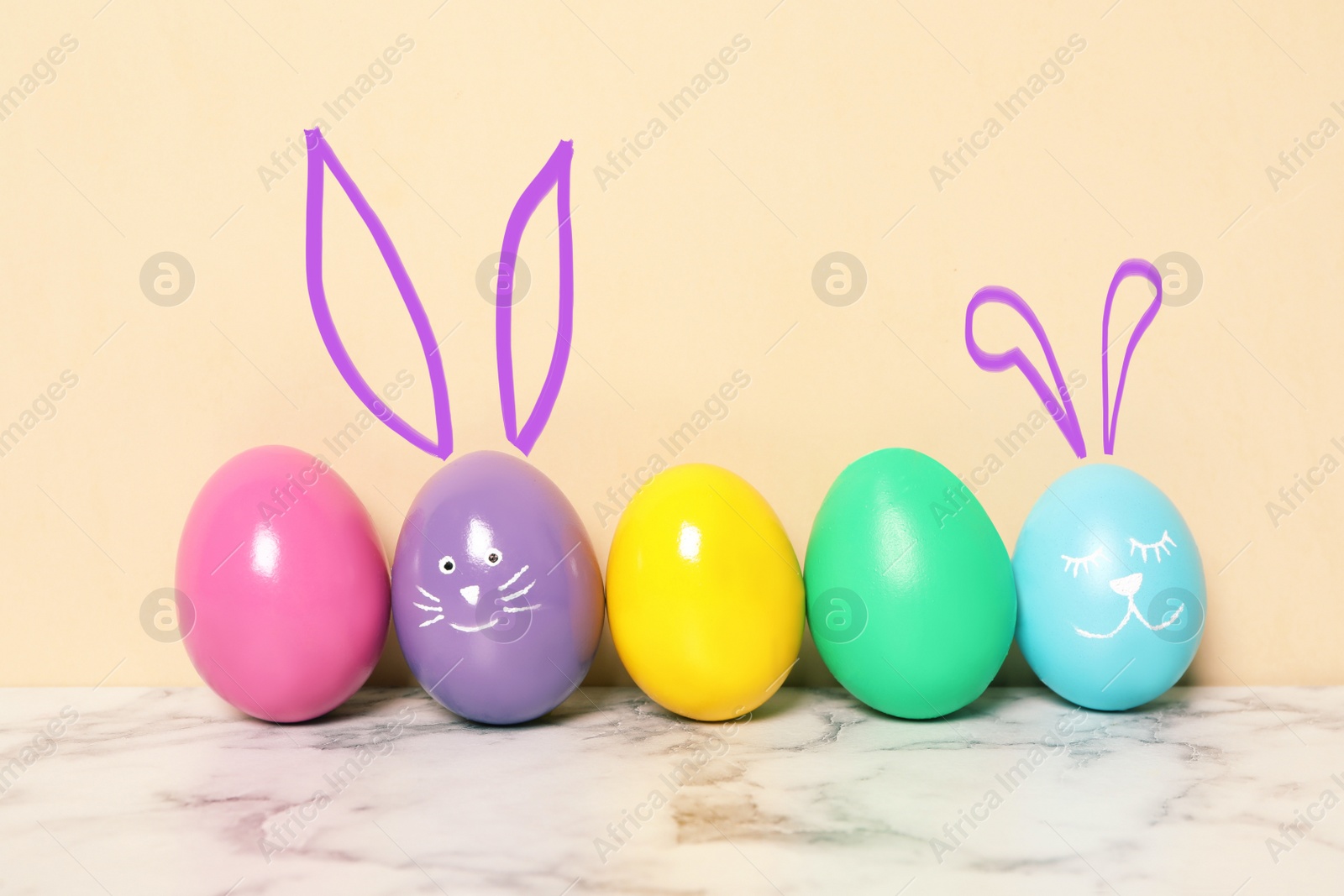 Image of Two eggs with drawn faces and ears as Easter bunnies among others on white marble table against beige background