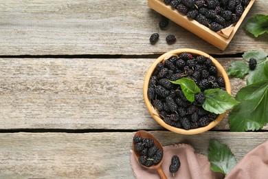 Bowl, spoon and crate of delicious ripe black mulberries on wooden table, flat lay. Space for text