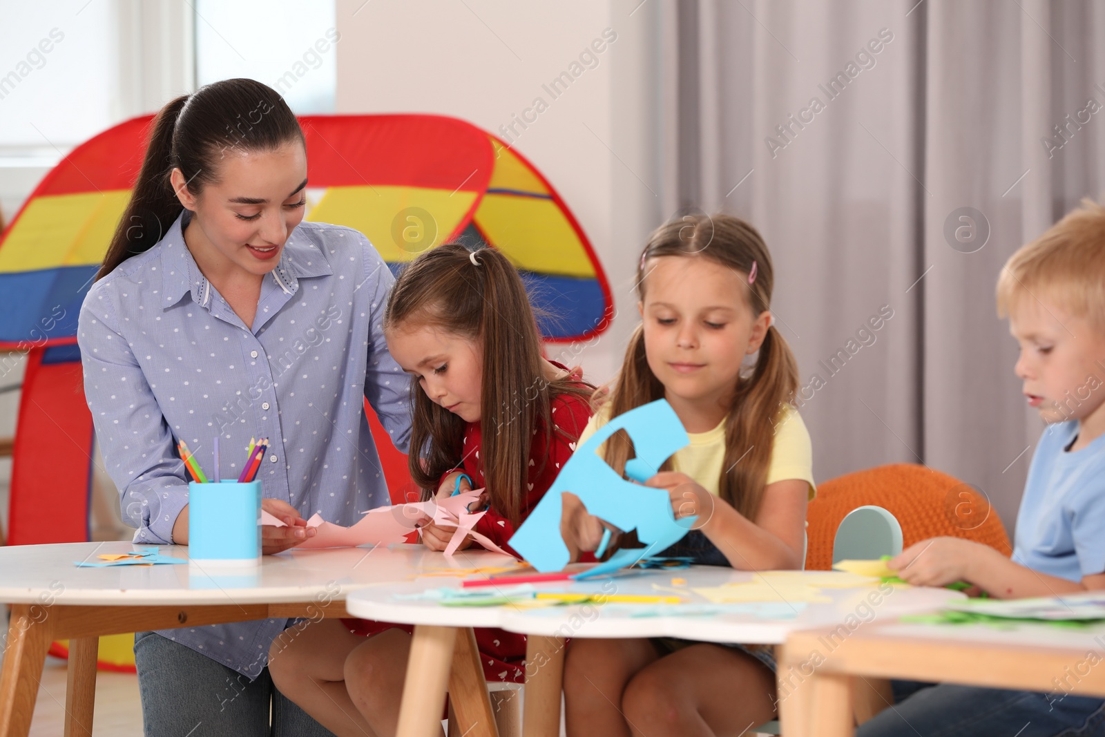 Photo of Nursery teacher and group of cute little children making toys from color paper at desks in kindergarten. Playtime activities