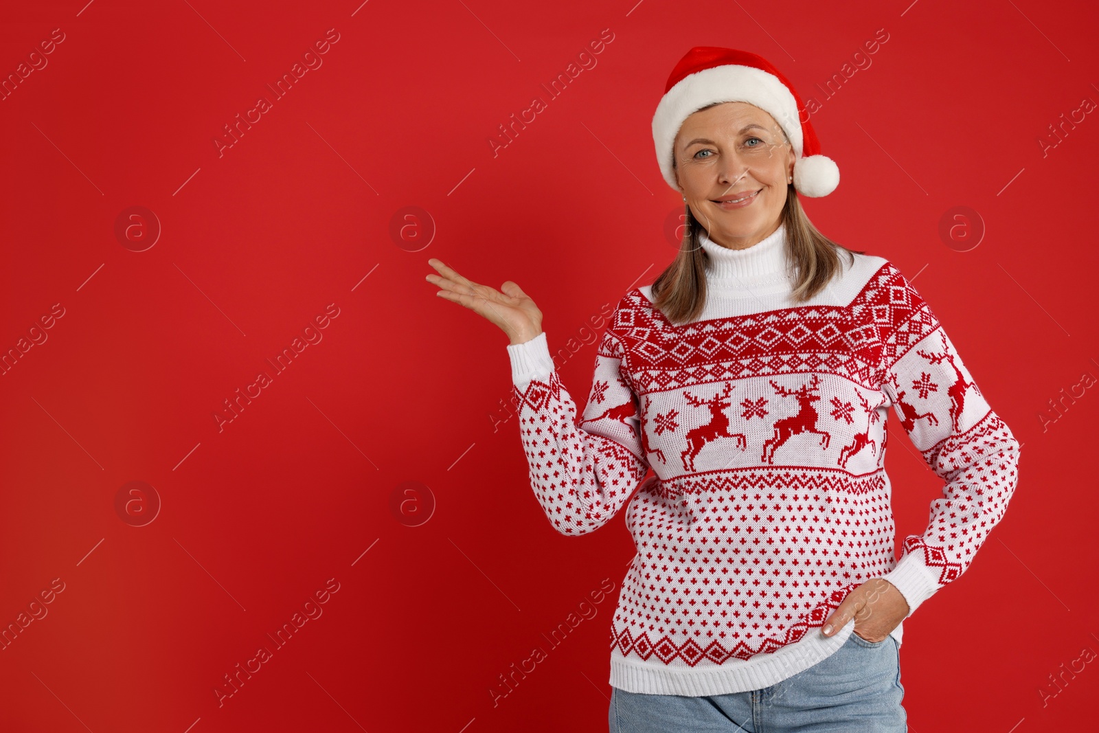 Photo of Happy senior woman in Christmas sweater and Santa hat showing something on red background. Space for text