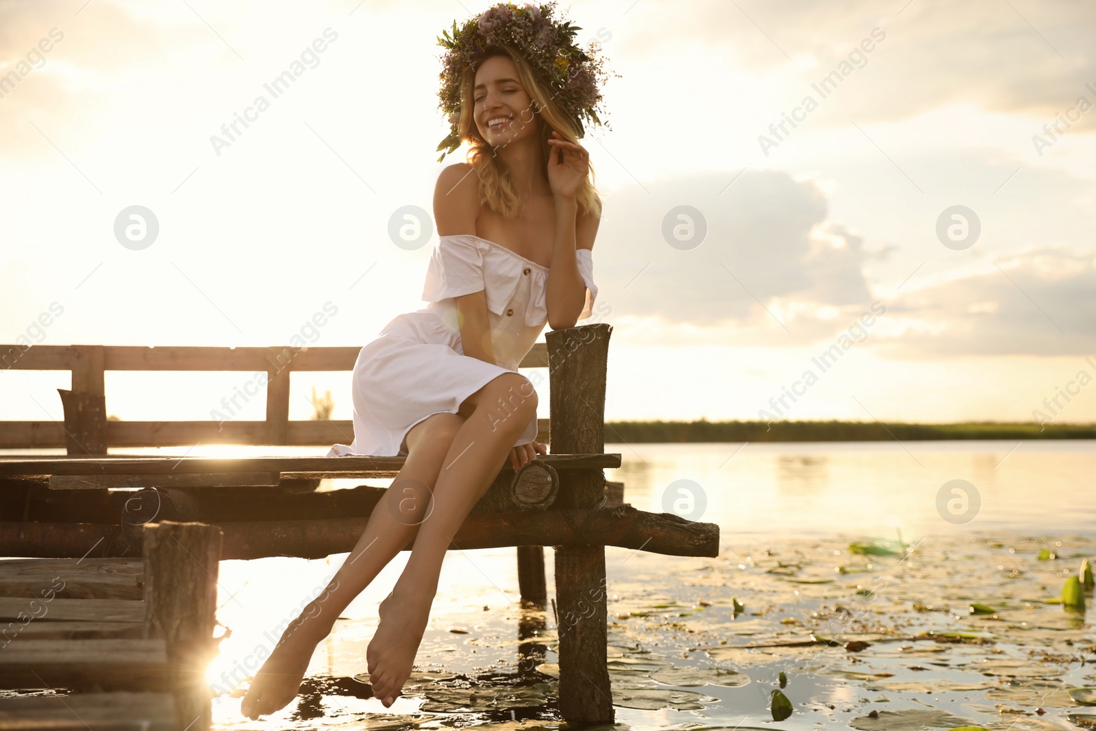 Photo of Young woman wearing wreath made of beautiful flowers on pier near river