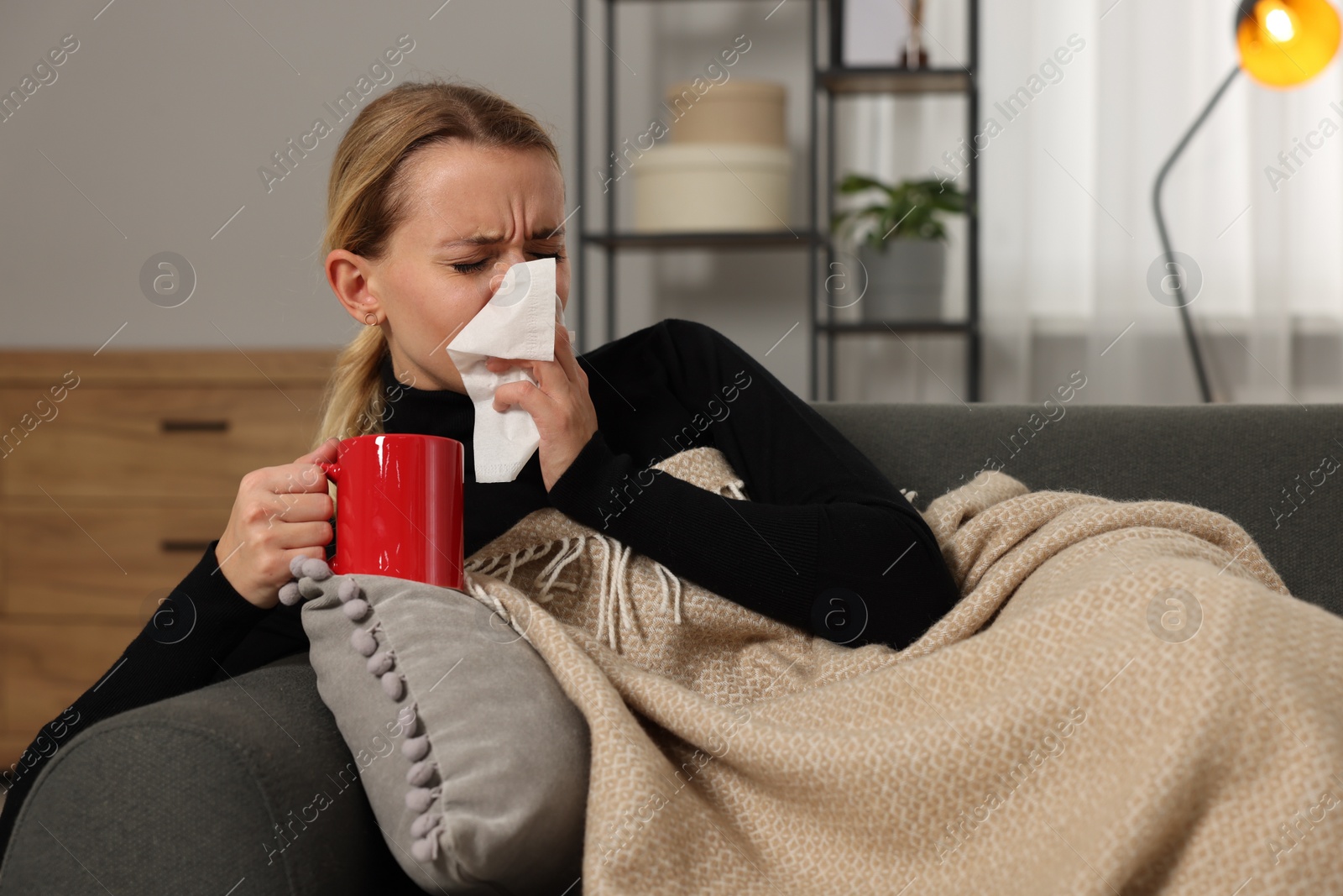 Photo of Sick woman with cup of drink blowing nose in tissue on sofa at home. Cold symptoms