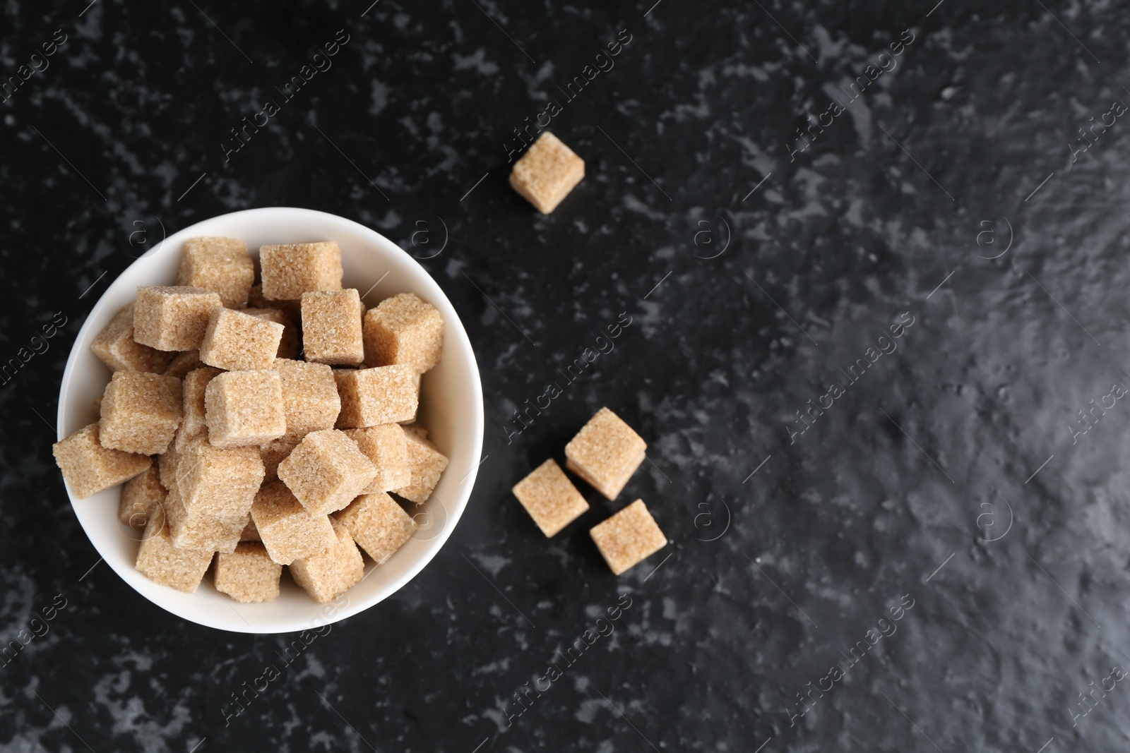 Photo of Brown sugar cubes in bowl on dark textured table, top view. Space for text