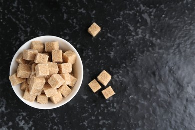 Photo of Brown sugar cubes in bowl on dark textured table, top view. Space for text