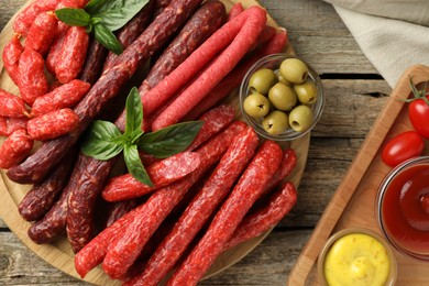 Different thin dry smoked sausages, basil, sauces and olives on wooden table, flat lay