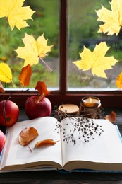 Photo of Book with dried flower, leaves as bookmark and ripe apples on table near window