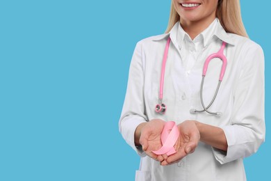 Photo of Doctor with stethoscope holding pink ribbon on light blue background, closeup and space for text. Breast cancer awareness