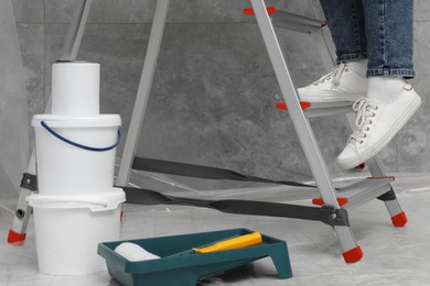 Photo of Woman climbing metallic folding ladder, tray with roller and buckets of paint indoors, closeup