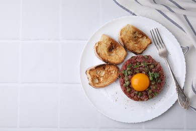 Photo of Tasty beef steak tartare served with yolk, capers, toasted bread and greens on white tiled table, top view. Space for text