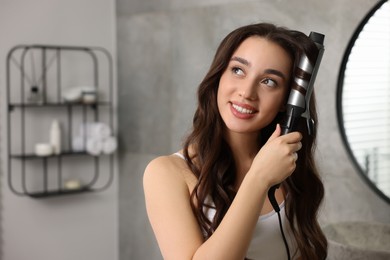 Photo of Smiling woman using curling hair iron in bathroom. Space for text