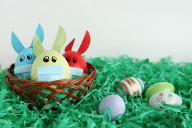 Photo of Painted eggs decorated with bunny ears and protective masks on green paper grass, space for text. Easter holiday during COVID-19 quarantine