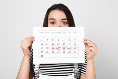 Young woman holding calendar with marked menstrual cycle days on light background