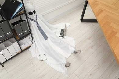 Overworked ghost. Man in white sheet with laptop on floor in office, above view
