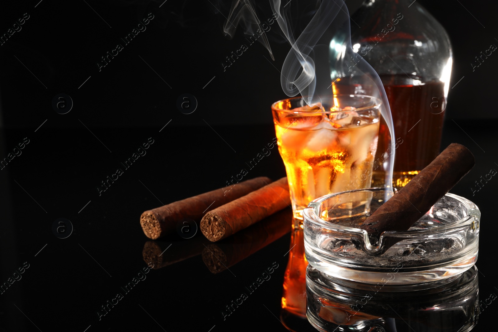 Photo of Smoldering cigar, ashtray and whiskey on black mirror surface. Space for text