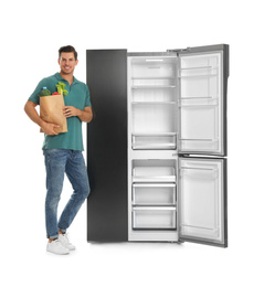 Man with bag of groceries near open empty refrigerator on white background