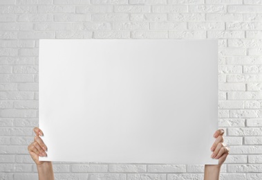 Woman holding blank poster near white brick wall, closeup. Mockup for design