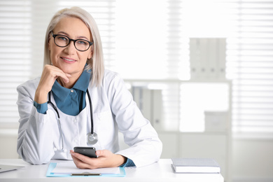 Mature female doctor with smartphone at table in office