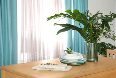 Dishware and plants on wooden table near window with elegant curtains