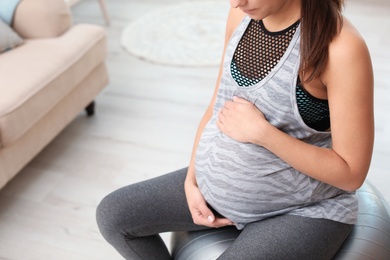 Photo of Young pregnant woman in fitness clothes sitting on exercise ball at home, closeup. Space for text