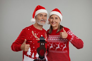 Photo of Senior couple in Christmas sweaters and Santa hats showing thumbs up on grey background